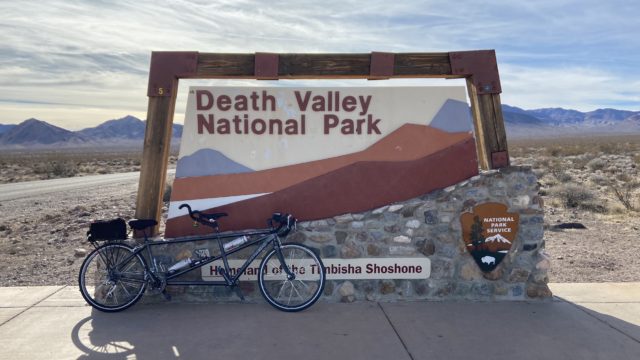 A gray tandem bike in front of a National Park Service sign for Death Valley National Park, the Homeland of the Timbisha Shoshone. The desert sprawls towards mountains in the background.