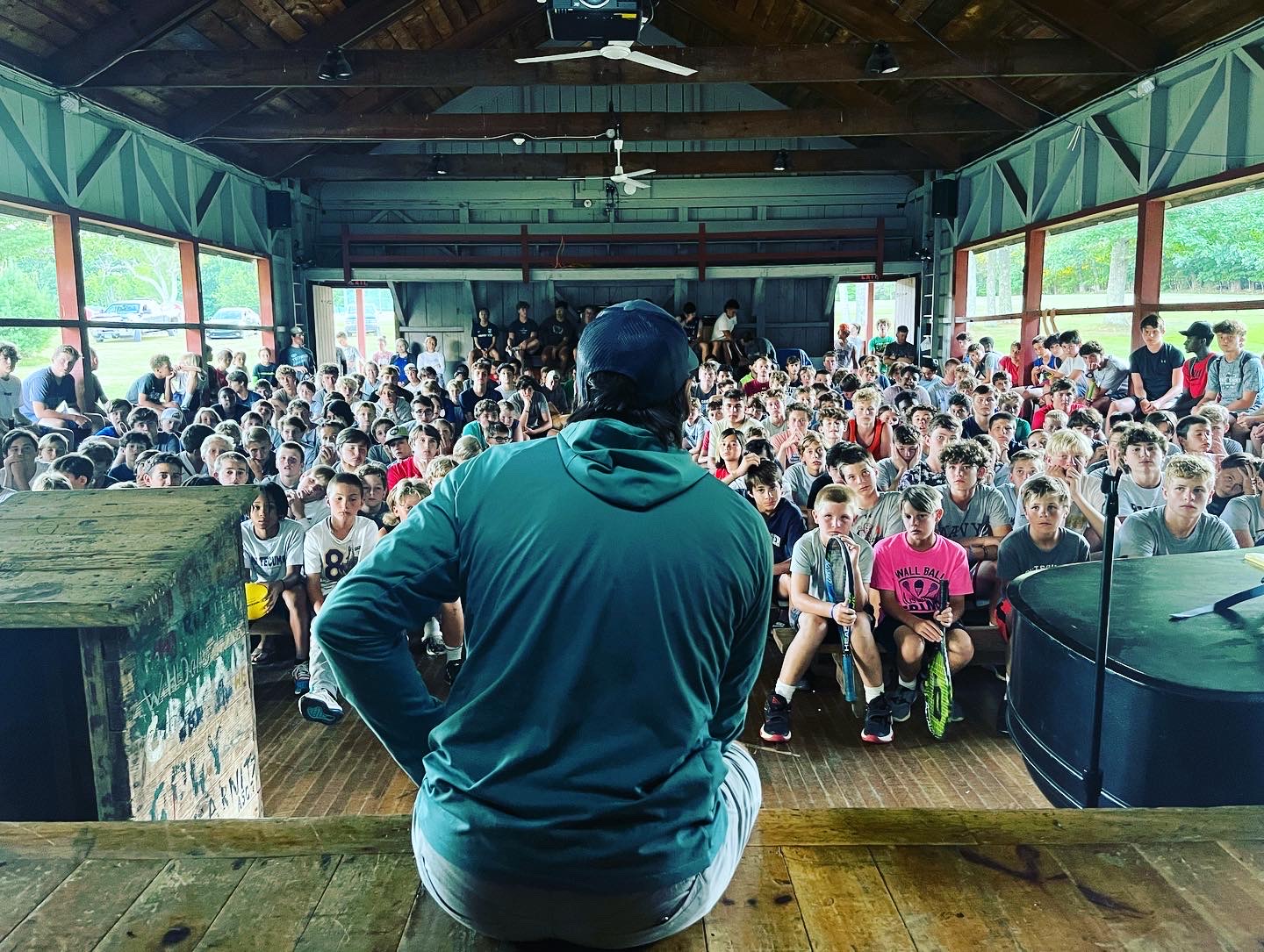 Erik speaking to a packed assembly hall. He is sitting on the stage wearing a green hooded sweatshirt with his back to the camera.