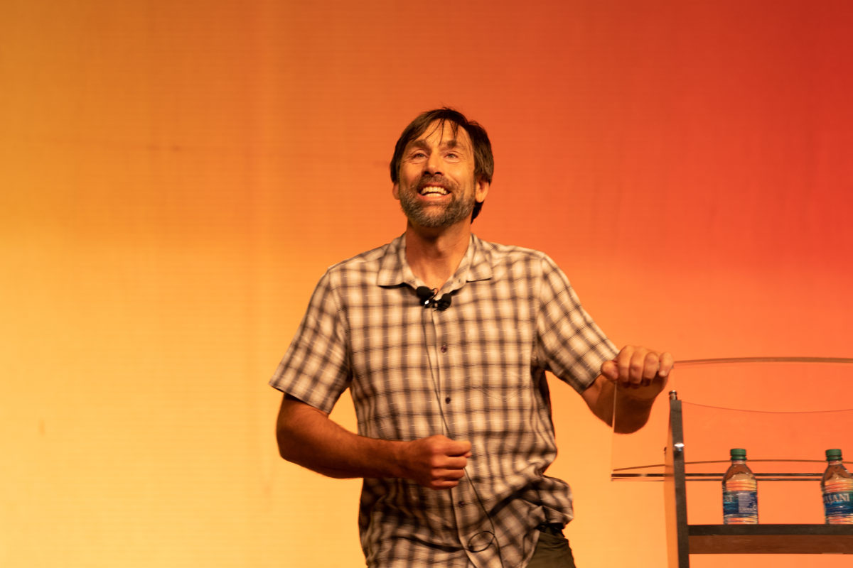Erik speaking on Saturday night. He is wearing a plaid shirt and smiling. He is holding the podium with his left hand and leaning in toward the audience.