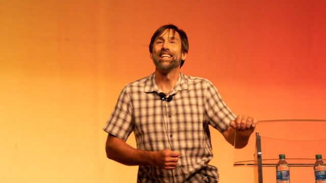 Erik speaking on Saturday night. He is wearing a plaid shirt and smiling. He is holding the podium with his left hand and leaning in toward the audience.